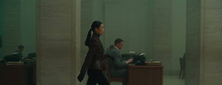A woman crosses a grey office between typewriters and answers the phone.