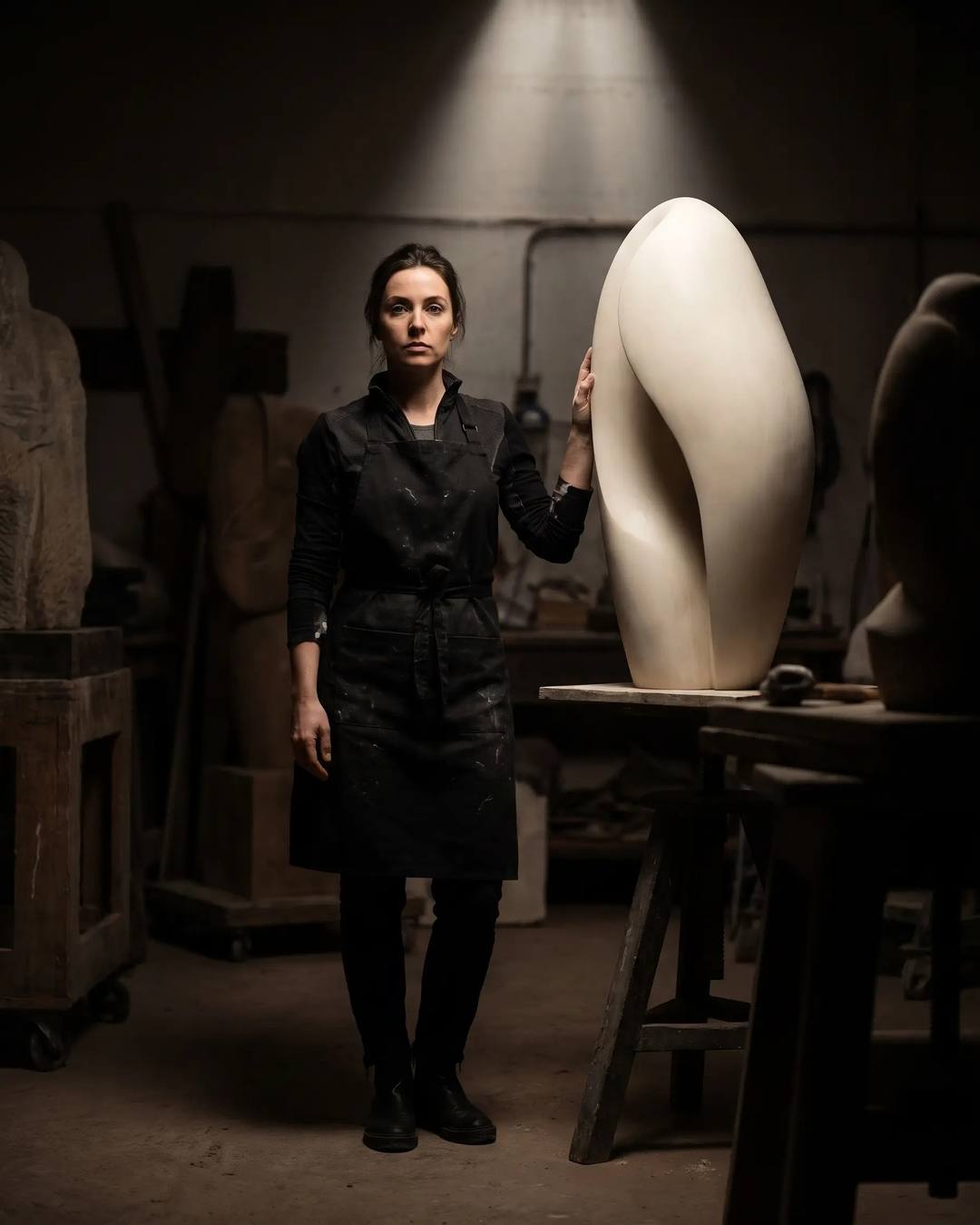 Una scultrice con il grembiule scuro appoggia la mano su un’unica scultura bianca.