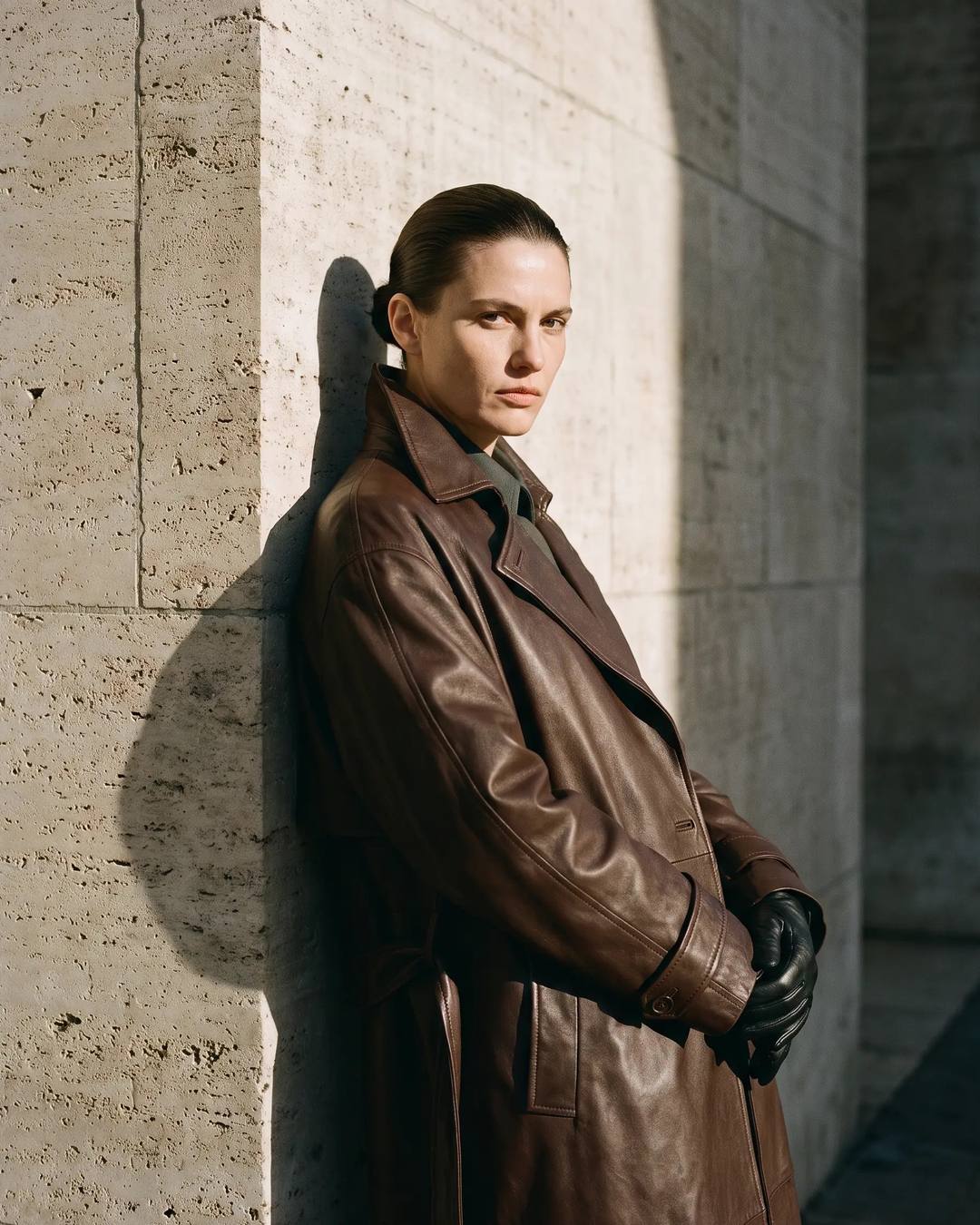 A woman in a brown leather trench coat leans against a travertine wall in hard afternoon sun.